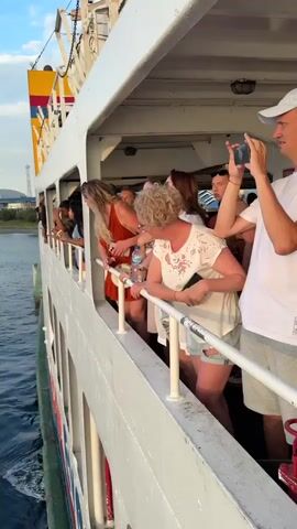 The video begins with a shot from the side of a ferry, looking out at the water. A group of people are standing on the deck, leaning over the railing, and looking out at the scenery. The water is calm and blue, and the sky is clear with a few scattered clouds. The ferry appears to be moving, as indicated by the wake in the water. The camera then pans to show the open sea with a distant island or landmass on the horizon. The sun is setting, casting a warm glow over the water and sky. Another ferry can be seen in the distance, moving across the water. The video ends with a close-up shot of the sunset, with the sun partially obscured by clouds.