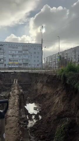 This is a video of a large excavation site in Usinsk, Russia. The scene features a deep trench dug into the earth, revealing layers of soil and some exposed pipes. The trench is partially filled with muddy water, indicating recent rain or groundwater.

In the background, there are several multi-story apartment buildings typical of Soviet-era architecture. A street runs along the edge of the excavation, with a sidewalk and a fence separating it from the construction area. Streetlights and utility poles are visible, suggesting an urban or suburban setting. A pedestrian crossing sign is also visible in the distance.

The weather appears to be overcast with dramatic, puffy clouds. The sun is partially visible through the clouds, casting a diffused light on the scene. The overall atmosphere is one of ongoing construction or repair work. No people are clearly visible in the frame, and the focus is on the excavation and its surroundings. The time of day appears to be daytime, likely late afternoon given the angle of the light. The activity taking place is the digging and exposing of underground utilities, likely for maintenance or replacement. There is no discernible text visible in the video.