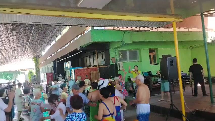A group of elderly people are dancing under a covered outdoor area. Some are wearing swimwear. There is a DJ booth with speakers and a person operating the equipment. Some people are holding up phones, possibly recording the event. The environment appears to be a resort or a community center with a casual atmosphere. The overall mood seems festive and joyful.