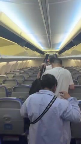 The video shows the interior of an airplane cabin. Multiple rows of empty seats with blue and gray upholstery are visible. Three people are walking down the aisle towards the front of the plane. The person in the foreground is wearing a light-colored shirt and has a strap from a bag across their chest. The person in the middle is wearing a white t-shirt and carrying a black backpack. The person at the front is wearing dark clothing. The cabin lighting is a cool blue, illuminating the overhead compartments and the curved ceiling.