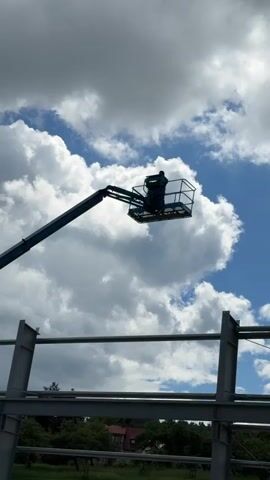 A person stands in the bucket of a boom lift, which is extended upwards towards a sky filled with dramatic clouds. The boom lift's arm, painted blue, stretches diagonally across the frame. Below the boom lift, the skeletal structure of a building, made of gray steel beams, is visible. The ground below the building structure is lined with trees and a hint of a red-roofed building. The scene appears to be a construction site under a partly cloudy sky.
