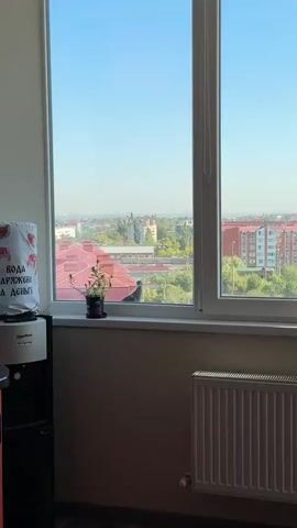 The video shows an indoor scene looking out a window. On the left, a water cooler is partially visible. On the windowsill, there is a potted plant and a pigeon perched on the sill. The pigeon is facing to the right. Outside the window, there is a view of buildings with red roofs, trees, and a distant cityscape under a clear blue sky. A white radiator is visible on the wall below the windowsill. The video is shot from a stationary position.