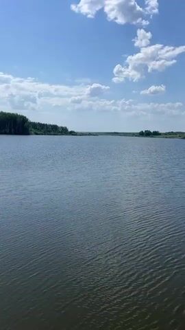 The video shows a body of water on a sunny day with a blue sky and scattered white clouds.  The first shot is a wide view of the lake or large pond.  In the distance, a line of trees borders the water.  A handrail is visible in the foreground. The next shot shows a metal structure over the water.  It has a bridge-like walkway and metal bars surrounding a hole in the water, possibly a drain.  The water around the structure is green.  The structure seems old and possibly in disrepair.