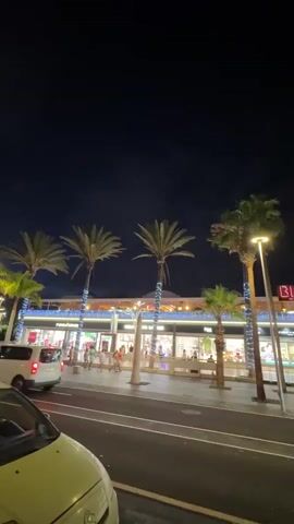 A nighttime street view of a shopping plaza. Palm trees with lights wrapped around their trunks line a sidewalk in front of shops. The shops have illuminated displays. People walk on the sidewalk. Cars are parked and driving on the street. The sky is dark blue and the overall scene is illuminated by artificial lights.