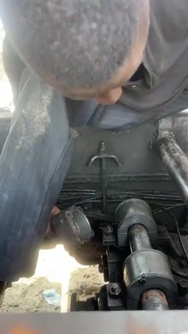 The video shows a close-up, downward-facing view of a person's head and torso as they lean over a mechanical engine. The person is wearing a dark shirt and grey pants. They appear to be working on the engine, which is located in what looks like an outdoor, dusty environment. A metal component with a handle is visible in the center of the frame, attached to the engine. The engine itself is greasy and appears to be part of a larger machine, possibly a vehicle. The lighting suggests it is daytime.
