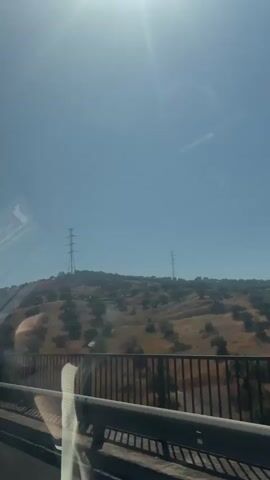 The video is taken from a vehicle moving along a road, likely a highway, judging by the guardrail in the foreground. The camera is positioned at a slight angle, capturing a view of a hilly landscape on the right. The sky is clear and blue, with the sun creating a lens flare effect near the top left of the frame. The hills are covered with sparse vegetation, appearing dry and brownish-yellow, with scattered dark green trees, possibly olive trees, dotting the slopes. Two tall power line towers are visible on the hilltop, one more prominent than the other. In the middle of the video, a blue road sign with white text and symbols appears. The text "1250 m" is at the top, followed by an arrow pointing right and the number "249". Below that, it reads "vía de servicio" (service road in Spanish) and shows icons for a car, a truck, and a tent. The vehicle continues to move, and the sign passes out of view, revealing more of the hilly terrain. The video captures a daytime scene with bright sunlight.