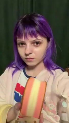 A person with purple hair is facing the camera and eating a multi-layered sandwich cake. The cake has three distinct layers: an outer layer of orange, a middle layer of white, and another layer of orange. The cake is partially wrapped in packaging with cartoon graphics. The person takes a bite of the cake, chews, and glances to the side. The background is a blurred green curtain. The person wears a peach-colored jacket over a shirt with red and blue graphics.