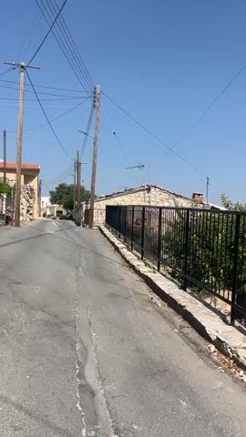 The video shows a view from a vehicle traveling down a narrow, paved street in what appears to be a village. The street is lined with stone buildings and walls, some of which are in disrepair. There are utility poles with numerous electrical wires running overhead. The sky is clear and blue, indicating a sunny day. In the distance, there are a few parked cars and some trees. The perspective suggests the vehicle is moving forward, creating a sense of motion.