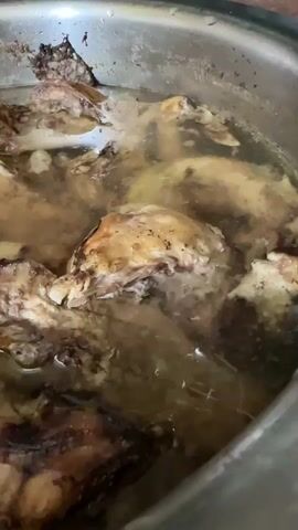 A close-up, overhead shot shows several pieces of roasted chicken, some with skin and bones visible, submerged in a pot of broth. The broth is cloudy and light brown, with some darker specks floating on the surface. The chicken pieces are varied in size and shape, with some appearing to be legs and thighs. The pot itself is a silver metal pot with a slightly tarnished rim. The lighting is dim, creating a somewhat somber atmosphere. The camera is stationary, with no visible movement.