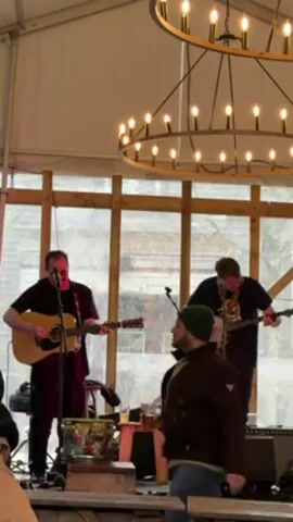 Two musicians are on a small stage, one playing an acoustic guitar and the other a banjo, inside what appears to be a tent-like structure.  A man wearing a green beanie and brown jacket walks by the stage in front of the band. The stage is simple, with wooden supports and a couple of speakers.  A large chandelier hangs overhead. The weather outside the tent appears overcast.