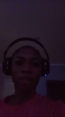 A close-up, shaky video shows a person wearing headphones and a pink shirt. The lighting is dim and changes colors throughout the video, shifting from a dark pink to blue. The background appears to be a wall with a door and some other objects that are not clearly visible. The person's face is mostly obscured by the low light.