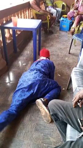 A man in a blue suit and red hat is lying on the floor, struggling to stand up and appears intoxicated. He is surrounded by people sitting on chairs around tables, some of whom are watching and laughing. The scene appears to be an outdoor gathering or celebration. The floor is tiled and there is a blue cooler and various drinks present.