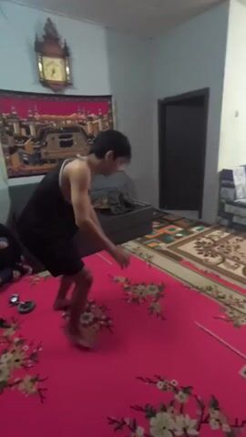 A young man in a black tank top and shorts is dancing barefoot on a bright pink rug with floral patterns. He's in a room with white walls and a couple of couches, one gray and one with blue and white checked fabric. There's a second rug beneath the couches that is tan with brown borders. There is a dark door in the background. The man looks like he is laughing and making exaggerated movements. There is a clock on the wall.