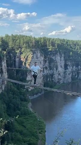 A woman stands on a rope bridge suspended over a river canyon. She is wearing a white t-shirt, black leggings, and a harness. She smiles and waves at the camera. The bridge is made of wooden planks and rope, and it spans a wide gap between two rocky cliffs. The cliffs are covered with trees, and the sky is blue with white clouds. The video is filmed from a slightly elevated perspective, looking down at the woman and the bridge.
