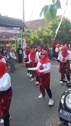 The video shows a group of women in red hijabs and red patterned skirts performing a dance. They are wearing white tops and white sneakers. The women are in a street with a crowd of people watching them. There are buildings and trees in the background. A motorcycle is parked in the foreground. The women appear to be participating in a parade or festival.