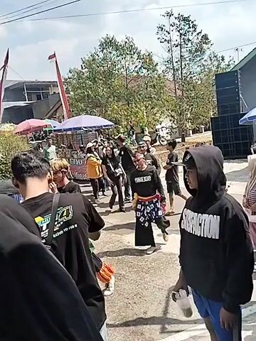 The video shows an outdoor gathering with a group of people. In the foreground, a person wearing a black hooded garment is partially visible, facing away from the camera. To their left, another person in a black t-shirt with white lettering is also facing away. Further into the scene, several individuals are walking and interacting. One person is wearing a black t-shirt with "NEVER GIVE UP" printed on it and a red and white headband, along with a sarong-like garment. Another individual in the background is wearing a yellow jacket. There are tents or stalls set up, possibly for vendors, with umbrellas providing shade. Trees and some buildings are visible in the background, suggesting a rural or semi-urban environment. A flagpole with flags is also present. The ground appears to be a paved surface. The overall atmosphere suggests a festive or community event.