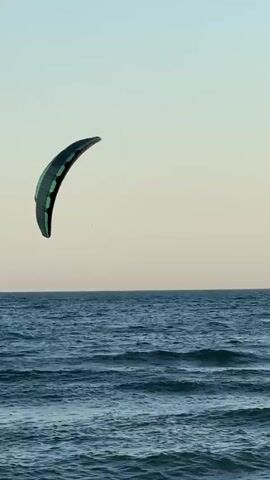 A kiteboarder is on the water. The kite is in the air. The sky is clear and blue. The water is blue and choppy. The kiteboarder is on a white board and is wearing dark clothing. The kite is green and black.
