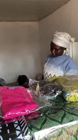 A woman wearing a light blue sweater with a white zig-zag pattern and a silver headscarf holds up a pair of burgundy platform sandals. She is standing behind a table laden with clothing items wrapped in plastic. The room appears to be a small shop or storage area, with plain walls and a white refrigerator visible in the background. The woman is facing the viewer and appears to be presenting the sandals.
