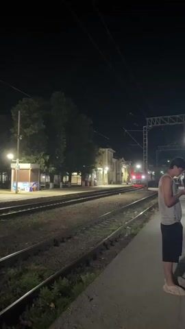 A train is approaching a dimly lit train station at night. A person stands on the platform looking at their phone. The train has its headlights on and is red and white. There are trees and a small building on the platform.