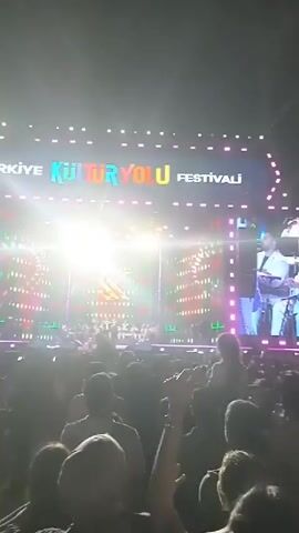 The video captures a live performance at a festival, likely a music event. A large stage displays the words "Kultur Yolu Festivali," indicating the name of the festival. On the stage, musicians perform, and their image is projected onto a large screen above them. A sizable crowd is visible in front of the stage, with many people raising their hands and cheering. The scene is brightly lit with stage lights, and the overall atmosphere appears energetic and lively.