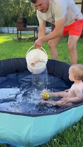 A man in a grey t-shirt and orange shorts pours water from a white bucket into a child's inflatable pool. A baby sits in the pool, facing away from the camera. The baby is naked and playing with a yellow and green toy. A small toy boat floats in the pool. The pool is set up on a grassy backyard. A grill and a wooden fence are visible in the background.