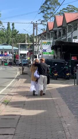 A couple walks arm-in-arm down a sidewalk on a sunny day. The man wears a dark long-sleeved shirt and light-colored pants. The woman wears a light-colored outfit. Behind them, shops line the street, with several motorcycles and a tuk-tuk parked along the curb. Trees are visible in the background.