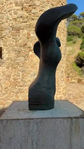 The video begins with a close-up shot of a dark, abstract sculpture positioned on a marble pedestal. The sculpture has a curved, organic form. In the background, a rustic stone tower stands prominently. The tower's surface is rough and uneven, constructed from a variety of stones. A small, barred window is visible on the tower's facade. To the right of the tower, a lush, green hillside slopes upwards, dotted with trees. The sky above is a clear, bright blue. The camera then pans upwards, revealing more of the stone tower, which appears to be quite tall. The top of the tower is covered with a white netting or fabric. The scene transitions to a lower angle, showing the underside of the netting on the tower's cap. The video then shifts focus to the sky above the tower, where several birds are seen flying. Finally, the view widens to encompass a white building with a pitched roof and two open doorways, set against the vast blue sky.