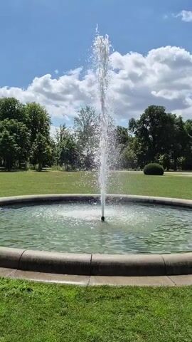 This is a vertical, eye-level shot of a fountain in a park on a sunny day. The fountain is in the foreground, with a strong jet of water shooting straight up into the air, creating a spray that catches the sunlight. The fountain is surrounded by a circular stone basin, which is filled with green water. Beyond the fountain, a well-maintained lawn stretches out, dotted with trees. In the distance, more trees form a dense line, suggesting a wooded area or the edge of a larger park. The sky above is a bright blue, with fluffy white clouds scattered across it. The overall impression is one of a peaceful, summery day in a park. There is no visible text in this media file.