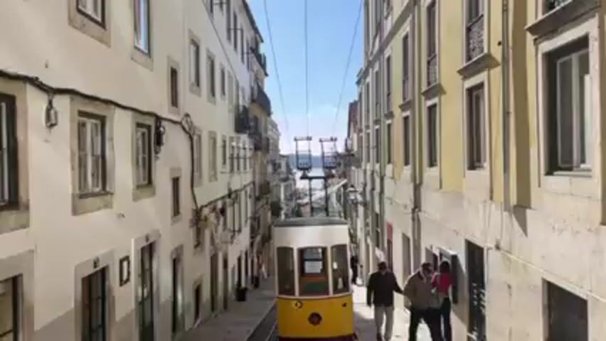 A yellow and white funicular is seen traveling down a narrow street in Lisbon, Portugal. Buildings on either side of the street are tall and made of stone.  The funicular travels down a set of tracks. People are walking on the sidewalk next to the funicular.  The sky is blue.  In the distance, the water is visible.