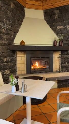 The camera pans slowly across a cozy restaurant setting featuring a stone fireplace with a roaring fire. In the foreground, there's a table set with a small vase of flowers, a pepper grinder, and a metal creamer, next to a cushioned bench along a stone wall. The floor is tiled in terracotta. The fireplace is accented with brick and wooden details, and the overall atmosphere suggests warmth and relaxation. In the background, indistinct voices can be heard.