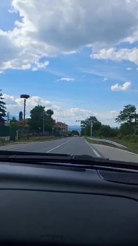 The video shows a view from inside a car looking out at a road on a sunny day. The sky is blue with white clouds. On the left side of the road, there are houses, trees, and a tall pole with a large nest on top. There are a few cars on the road ahead. On the right side of the road, there are trees and a white barrier. A traffic light or camera system is visible over the road. The dashboard of the car is visible in the foreground.