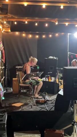 A blonde person with their hair pulled back in a bun, wearing a patterned tank top and green shorts, is playing a keyboard while sitting on a chair on a small stage. The person's feet are bare, and they are wearing sandals. The stage is lit by string lights and a spotlight. There are musical instruments and equipment on and around the stage, including speakers, a guitar, and what appears to be a cajon drum. Another person with a beard is visible in the background on the right side of the stage. The scene appears to be an indoor performance space.