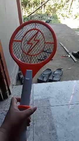 A first-person perspective shot shows a person holding an orange and white electric mosquito swatter. The swatter has a circular, netted head with a lightning bolt symbol in the center. The handle is white with an orange button and a red cap. The background is blurry but appears to be an outdoor setting with a wall, some greenery, and a few pairs of flip-flops on the ground. The person's hand is visible holding the swatter, and the swatter is moved slightly up and down.