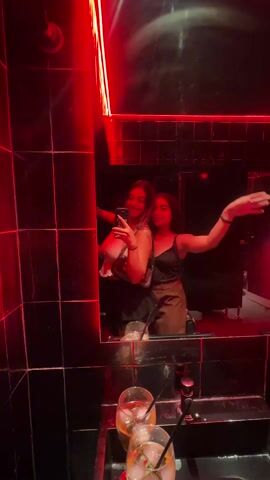 Two young women are taking a selfie in a bathroom with red lighting. The bathroom has dark tile and a large mirror. Two cocktails are sitting on the countertop. One woman is holding a smartphone and taking a picture. Both women are smiling and posing for the camera. The music playing in the background is loud.