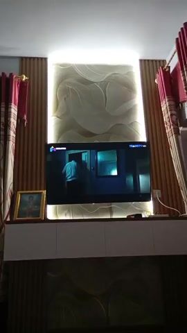 A flat-screen television is mounted on a white cabinet. The television is displaying a scene from a show where a person wearing an orange shirt and dark pants is walking through a doorway. To the left of the television, there is a framed picture. The wall behind the television is decorated with a large, light-colored wallpaper featuring abstract designs. On either side of the television, there are vertical wooden slats, and a red curtain is visible on the far left and right edges of the frame. The area above the television is illuminated by a strip of white LED lights. The cabinet beneath the television also has decorative elements on its front.