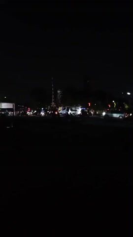 The video captures a dark outdoor scene, likely at night, illuminated by various artificial lights. In the center of the frame, a white van is visible with its headlights on, casting a bright glow. To its left and right, several motorcycles and a few cars are present, their lights also contributing to the illumination. A traffic signal or pole structure stands prominently behind the van. The overall impression is of a traffic intersection or a gathering of vehicles and people, with the darkness of the night contrasting with the artificial lights.