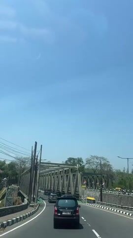 The video shows a view from inside a vehicle traveling on a road towards a steel truss bridge. The sky is clear and blue with a faint wispy cloud. On the left side of the road, there are utility poles and wires. The road has a white and black striped curb and a green painted bike lane. Several cars are visible on the road, including a black hatchback in the foreground and a dark SUV further ahead. The bridge is made of metal beams forming a lattice structure overhead, and the road continues over it. Trees and some buildings are visible beyond the bridge on both sides. The camera is moving forward, capturing the approach to and passage under the bridge.