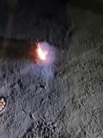 The video shows a firecracker exploding on a dark, possibly outdoor, surface. The explosion is captured in bright flashes of orange and yellow, accompanied by trails of sparks shooting outwards. Smoke billows upwards, partially obscuring the initial flash. There are no visible people or surrounding objects in the frame. The focus remains solely on the firecracker's explosion.