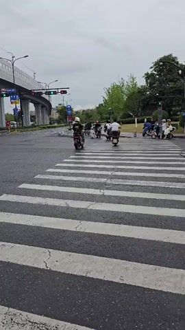 A group of people on scooters are crossing a street at a pedestrian crossing. One person in the foreground is wearing a blue raincoat. There is an overpass in the background, and traffic lights are visible. The sky is cloudy.