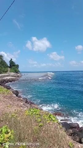 A video shows a view of the ocean from a rocky coastline. The water is a vibrant turquoise color, and waves are breaking on a coral reef that extends out into the sea. The sky is blue with scattered white clouds. There are some palm trees and vegetation along the coastline. The camera is positioned slightly above the ground, looking out towards the water. A power line is visible in the upper left corner of the frame. There are a few people visible near the water's edge on the left side of the frame. A watermark in the bottom right corner of the video indicates it was filmed on an Infinix Note 40 Pro. The video is very stable, suggesting it was filmed from a stationary position.