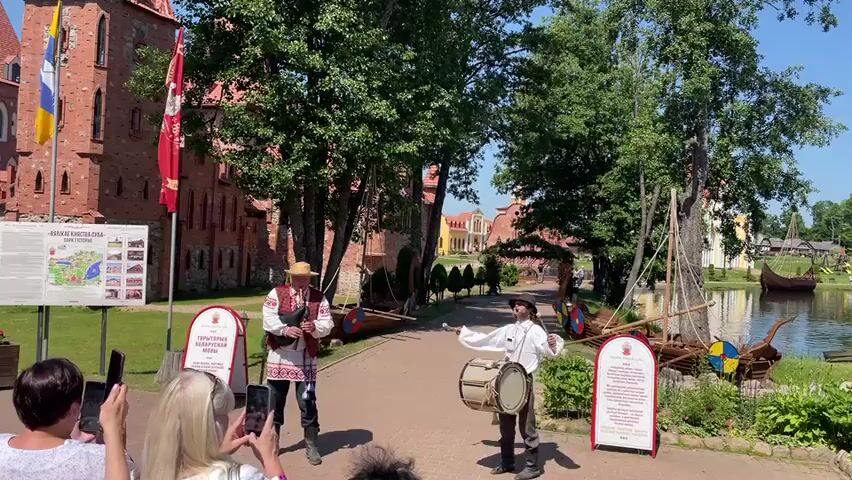 The video shows two men in historical costumes performing in front of a medieval-style castle. The man on the left wears a straw hat, a white shirt with red and black embroidery, and dark pants. He plays a flute or a similar wind instrument. The man on the right wears a hat, a white shirt, and dark pants, and plays a drum. They are positioned on a cobblestone path. On the left side of the path is a brick castle with flags. To the right of the path is a body of water with a Viking-style boat and other historical structures. Several people are in the foreground, watching the performance and taking pictures with their phones. There are also informational signs on the left and right sides of the path. The environment appears to be outdoors on a sunny day with green trees and grass.