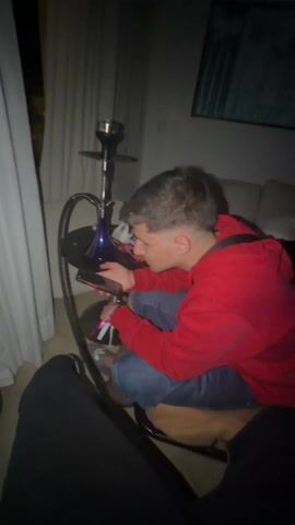 A young man in a red hoodie is fiddling with something in his hands next to a hookah on a small, round table. He's kneeling on a brown cushion. The camera moves to show another person lying on a bed behind him, then comes back to focus on the first man, who is now looking directly at the camera. There is a picture hanging on the wall behind him, and a window with white curtains to his left. The room is dimly lit.