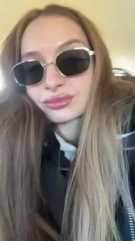 A woman with long brown hair and sunglasses is looking at the camera with a slight smirk. She is wearing a black jacket and white shirt. The background appears to be indoors and slightly blurry. Music is playing in the background.