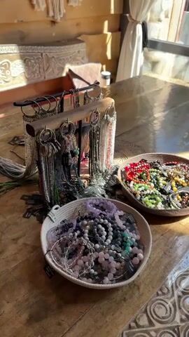 The scene shows a wooden table displaying jewelry.  There are two round, shallow baskets containing beaded bracelets in a variety of colors including purple, pink, blue, green, red, yellow, and orange.  Behind the baskets is a metal display stand with hanging earrings and necklaces of various styles and colors.  There are small black price tags near the items. The table has a textured, almost mosaic surface. Sunlight streams in through a window and reflects off the table surface.  In the background, there is a cushioned bench and a white curtain hanging near the window. The overall setting suggests a craft fair or a boutique.