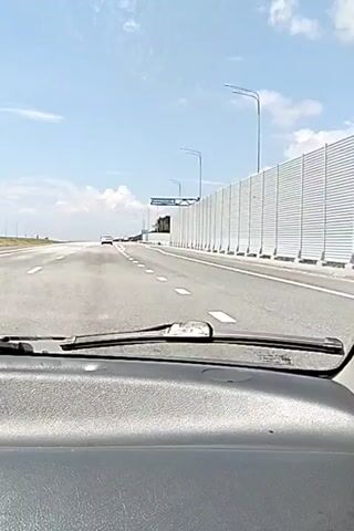 The video shows a first-person view from inside a car driving on a highway. The dashboard and windshield are visible in the foreground. The road is paved and has white lane markings. On the right side of the road, there is a tall, corrugated metal sound barrier. Beyond the barrier, there are trees and a blue sky with white clouds. There are a few other cars visible on the highway ahead. The video is taken during the daytime under sunny conditions. The car appears to be moving at a moderate speed. The camera captures a brief view of the side mirror and the side window, showing the road and the sound barrier. The landscape suggests a suburban or exurban environment.