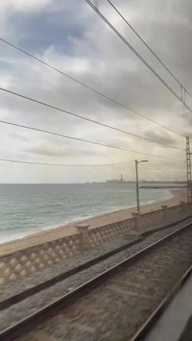The video captures a view from a moving train, looking out a window. In the foreground, train tracks run parallel to a textured, light-colored concrete or masonry wall. Beyond the wall is a sandy beach leading to an ocean with light blue-green water. The sky is overcast with patches of brighter light. Overhead electrical lines and support poles are visible. In the distance, a city skyline or industrial port area with buildings and cranes can be seen along the coastline. Sparse vegetation is scattered along the beach between the wall and the distant city. The train is in motion, moving forward.