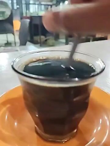 A person is shown stirring a cup of black coffee with a spoon. The coffee cup is a clear glass with an orange saucer. The table on which the coffee sits is white. The background shows an outdoor eating area, with tables, chairs and people walking. The lighting is dim, suggesting it is nighttime. After stirring, the spoon is left in the cup.