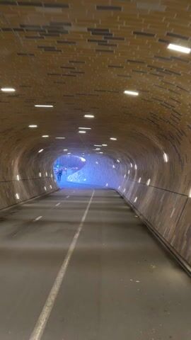 The video shows the interior of a tunnel. The tunnel has a rounded, brickwork ceiling with recessed lights. The tunnel road is asphalt with a solid white line down the middle. At the end of the tunnel, blurred figures of cyclists are visible in the daylight.