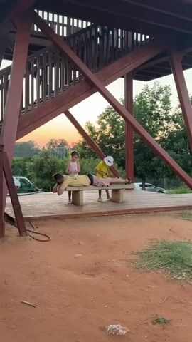 A man is lying face down on a park bench with his arms extended. A young girl stands to his left, looking down at him. To the man's right, a child wearing a yellow shirt and a white hat is sitting on the bench, looking down at the man. The scene takes place on a wooden platform with a large, reddish-brown wooden structure above. The background shows trees and a road with cars. The sky in the background is orange and pink, indicating it is sunset.