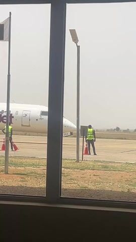 Here is a description of the video:
The video shows an airplane on a tarmac, seen through a window. The plane is a Value Jet plane, white with purple writing. Two men in yellow vests are standing near the plane, on either side of orange cones. A "No Smoking" sign and a Nigerian flag are visible in front of the window, along with a plant in a gray pot. The scene appears to be taking place at an airport.